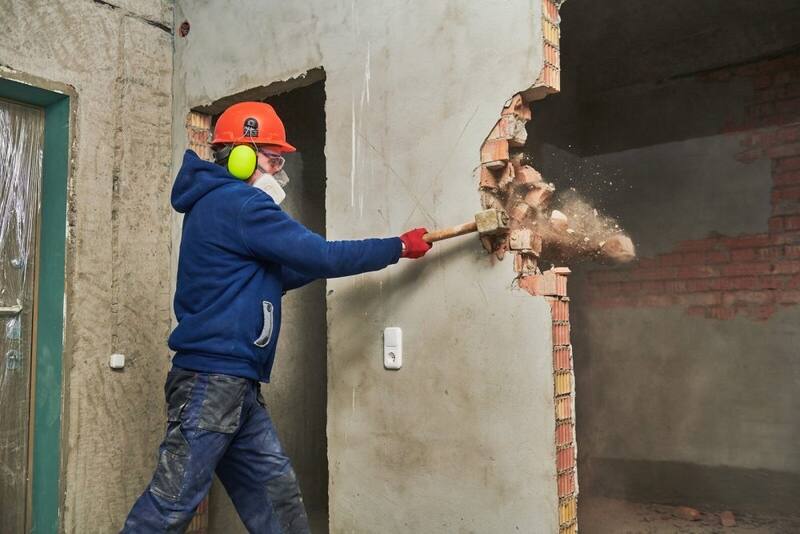 Pracovník v prilbe a s chráničmi sluchu búra tehlovú priečku kladivom